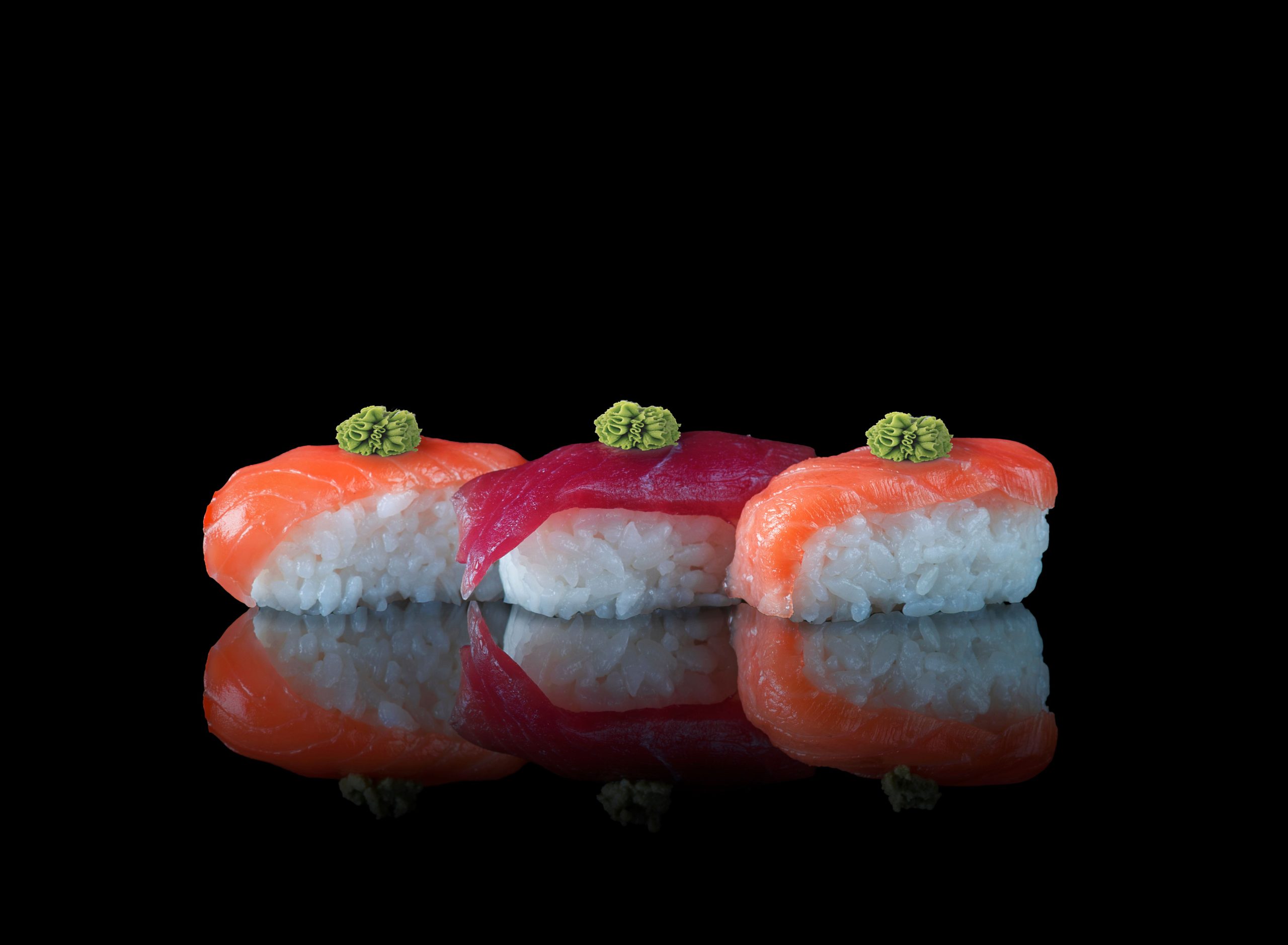 A close-up of nigiri sushi with salmon and tuna topped with wasabi on a reflective surface.